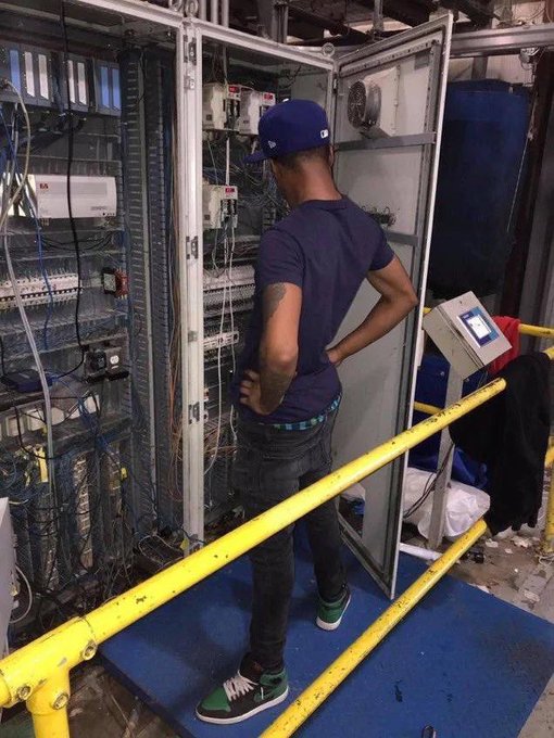 A man in a blue baseball cap, purple t-shirt, dark jeans, and green sneakers stands with his hands on his hips looking into an open industrial electrical control cabinet filled with wires.