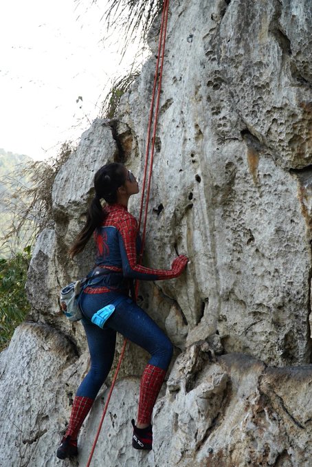 A person wearing a Spider-Man costume is rock climbing up a rugged, light-colored cliff face, secured by a red rope.