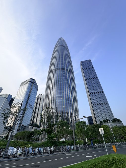 A tall, bullet-shaped skyscraper with a glass facade towers over surrounding modern buildings along a tree-lined street.