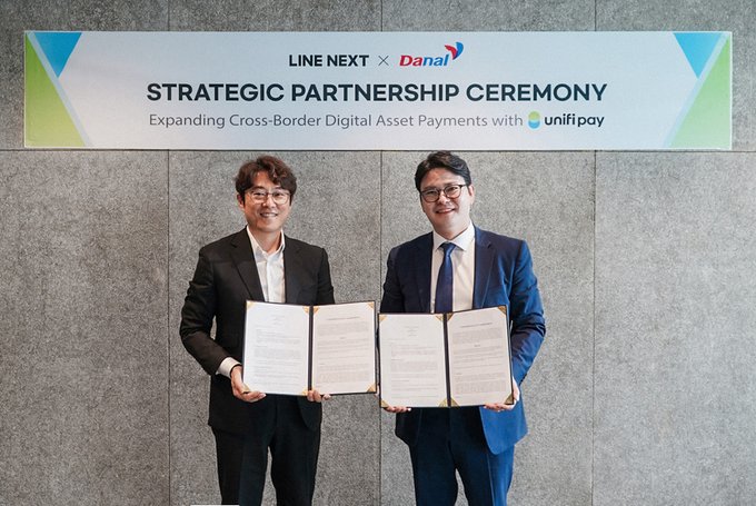 The image shows two men standing next to each other holding papers with their hands together. They are both smiling for the camera while posing for the photo. The background of the image features a sign that says "LINE NEXT Strategic Partnership Ceremony" and "Expanding Cross-Border Digital Assets Payments with Unifi". This suggests that the event is related to business partnerships or financial transactions, possibly involving the use of digital assets and cross-border payments.