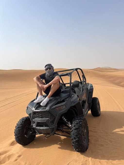 A man wearing sunglasses, a bandana, and casual athletic wear sitting on the hood of a gray Polaris RZR side-by-side UTV in a vast desert landscape with sand dunes.