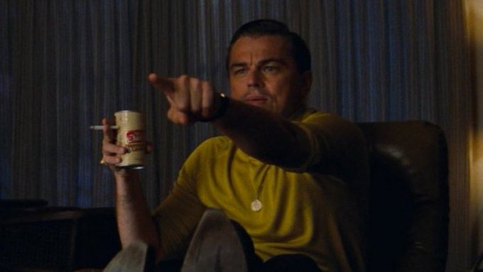 A man in a yellow shirt sits in a chair holding a beer can and cigarette, pointing directly at the camera in a meme-like gesture often used to imply suspicion or accusation. The caption connects this pointing pose to a playful "Arbitrum mentioned" reference, framing the gesture as a pointed callout to the cryptocurrency being discussed.