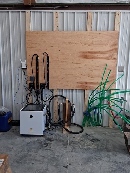 A workshop or garage space with a wooden plywood board mounted on a corrugated metal wall, featuring charging equipment, cables, and a white machine unit on the concrete floor, next to a bundle of green hoses.