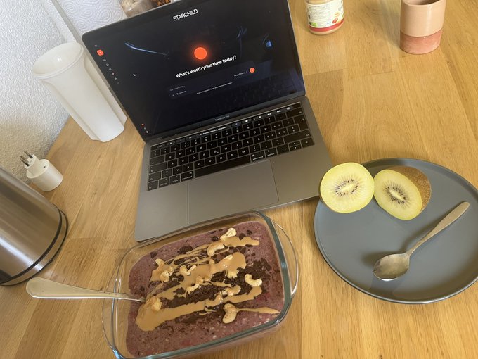 The image shows a laptop computer placed on top of a wooden table, with a plate of food containing a bowl of oatmeal topped with peanut butter and honey, as well as a kiwi slice and a spoon nearby. The background appears to be a kitchen setting with various items such as cups, bowls, and other utensils visible. The image also contains text that reads "AI builder breakfast".