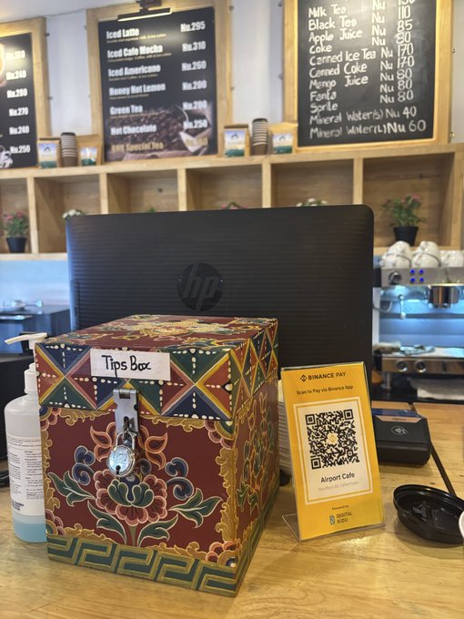A coffee shop counter with a menu board displaying items like "Iced Latte" and "Americano", an HP computer monitor, a decorative "Tips Box", and a Binance Pay QR code stand.