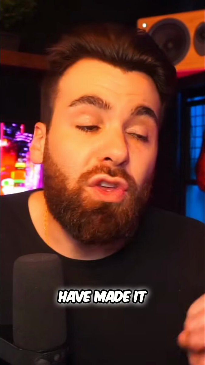 The image features a man with a beard wearing a black shirt, holding a microphone and speaking into it. There are two speakers visible behind him, one of which has a sticker that says "Have Made It" written on it. The background is dark, making the man stand out in the photo.