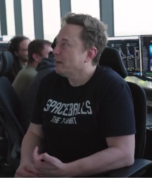 Elon Musk wearing a black 'SPACEBALLS THE T-SHIRT' t-shirt, sitting in what appears to be a mission control room with monitors in the background.