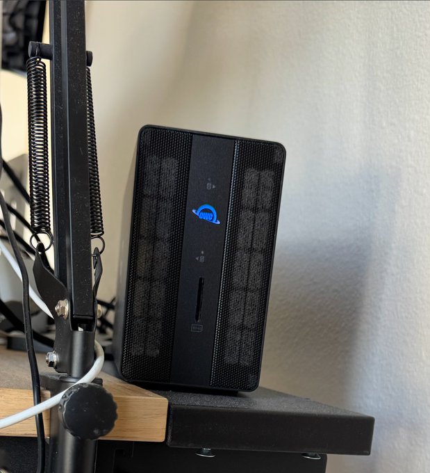 A black electronic device with a blue cloud logo sits on a desk next to a microphone stand with tension springs, appearing to be a hardware wallet or similar gadget.