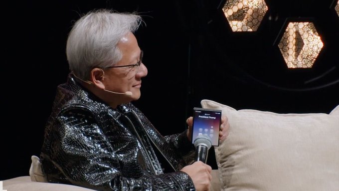 A side-profile photograph of an older man with gray hair and glasses, wearing a black sequined jacket and a headset microphone, holding a microphone and a smartphone during what appears to be a presentation or interview.