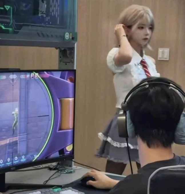 A scene from an internet café or gaming center showing a person wearing headphones playing a first-person shooter game on an AOC monitor, while a cosplayer dressed as a blonde anime-style character in a white blouse, red tie, and gray skirt stands in the background.