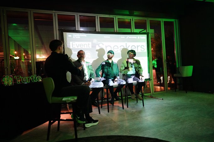 Four men sit on stools on a dimly lit stage with green lighting during an "Event Speakers" presentation at a Liquidy Berlin event.