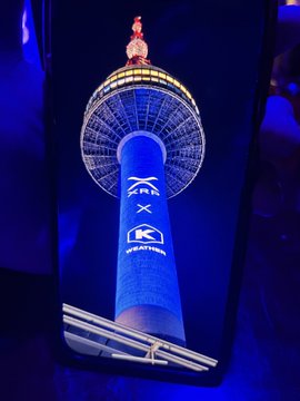 A smartphone displaying a photograph of a tall blue-lit tower with 'XR' and 'KC' signage, taken in a dark environment.