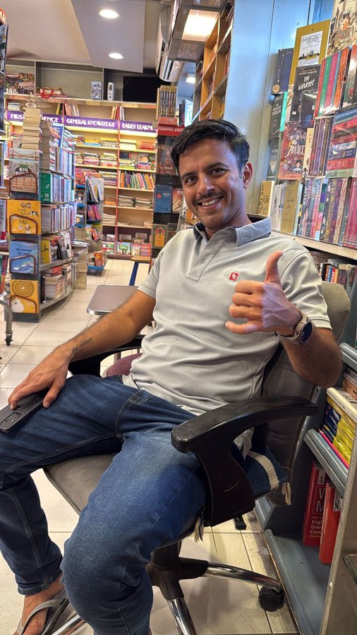 A man with a mustache smiling and giving a shaka sign while sitting in a chair inside a bookstore, surrounded by shelves of books.