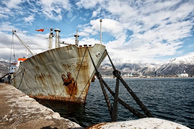 A large, rusted cargo ship is docked at a pier with thick mooring ropes, set against a backdrop of mountains and a cloudy blue sky.