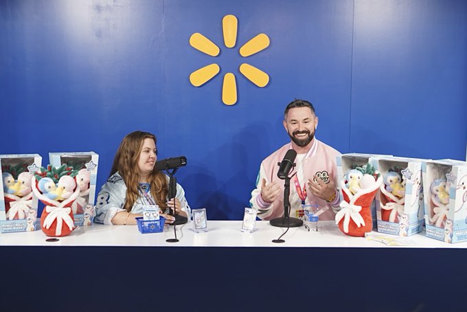 Two presenters sit at a table displaying boxed Pudgy Penguins plush toys in front of a blue backdrop featuring Walmart's yellow spark logo, appearing to host a segment for Walmart's official NY Comic Con livestream. The scene directly supports the post about Pudgy Penguins being featured on Walmart's Comic Con broadcast, showcasing the brand's plush merchandise to a wider audience.
