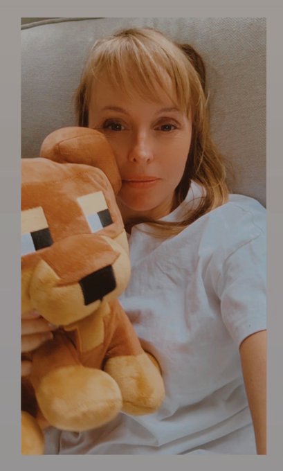 A Polaroid-style photo of a young blonde woman with bangs holding a Minecraft fox plush toy while wearing a white t-shirt.