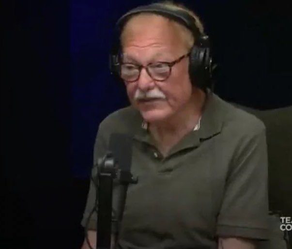 An older man with glasses, a white mustache, and black headphones wearing an olive green polo shirt, speaking into a studio microphone against a dark background.