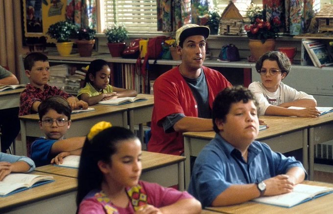 A classroom scene from a movie or TV show shows a teacher in a red shirt and baseball cap standing among students seated at their desks.