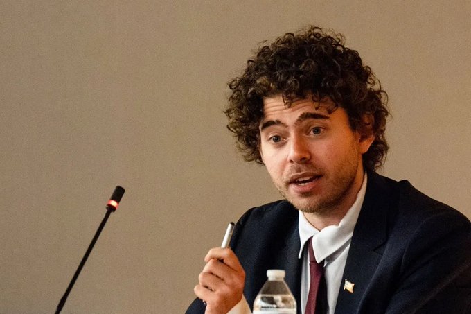 A young man with curly hair, wearing a dark suit and tie, speaks into a microphone while holding a pen, with a water bottle visible on the table before him. The image likely depicts a Polymarket spokesperson or executive addressing the reported credit card fraud and money laundering allegations mentioned in the post.