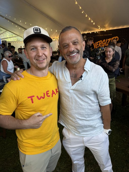 Two men smile and pose together at an outdoor event under a tent, one wearing a yellow 'TWEAK' t-shirt and the other in a white linen shirt, with a 'VERGIZZ' banner in the background.