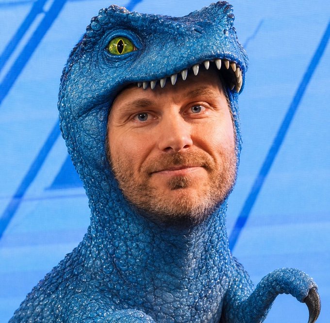 A man wearing a blue velociraptor costume headpiece featuring a yellow eye and white teeth, posing against a blue geometric background.