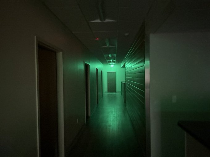 A dimly lit hallway is illuminated by eerie green emergency lighting, with an exit sign glowing at the far end near a closed door.