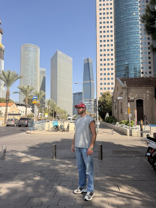 A man in a red cap and gray sleeveless shirt stands on a paved plaza in front of a row of towering modern skyscrapers, including glass high-rises and a stone church. The giveaway post invites followers to drop their Solana wallet addresses to win $250, with the urban skyline serving as a scenic backdrop that doesn't directly tie to the crypto giveaway theme.