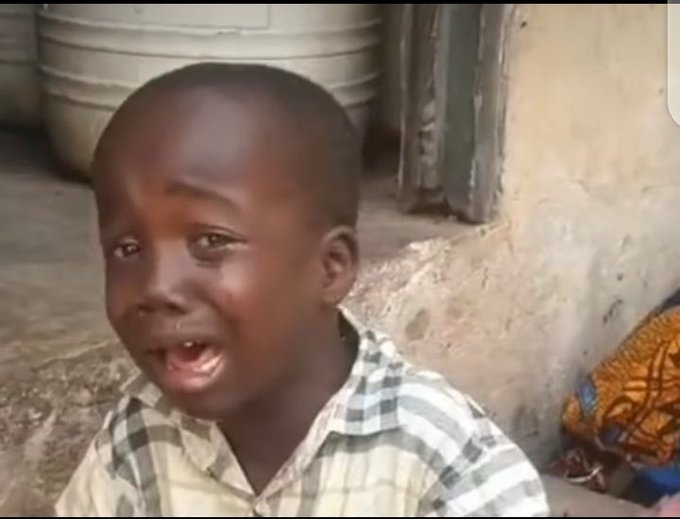A young boy in a plaid shirt appears to be crying while sitting on the ground near a large metal pot in what looks like a village setting.