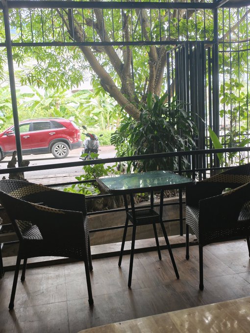 The image shows a balcony with two wicker chairs, one of which has a small table next to it. The chairs are positioned facing each other, creating an intimate seating area. A red car can be seen parked outside the building, adding color to the scene. In addition to the chairs and table, there is also a potted plant on the balcony, contributing to the overall ambiance of the space.