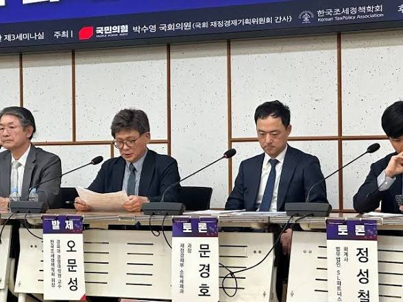 A panel of South Korean officials sits at a podium labeled "한국조세재정학회" (Korean Tax & Finance Association), with nameplates identifying speakers including 오태석, 민종학, and 정성천, as the image accompanies news of South Korea's opposition moving to scrap the planned 22% crypto tax set for 2027.