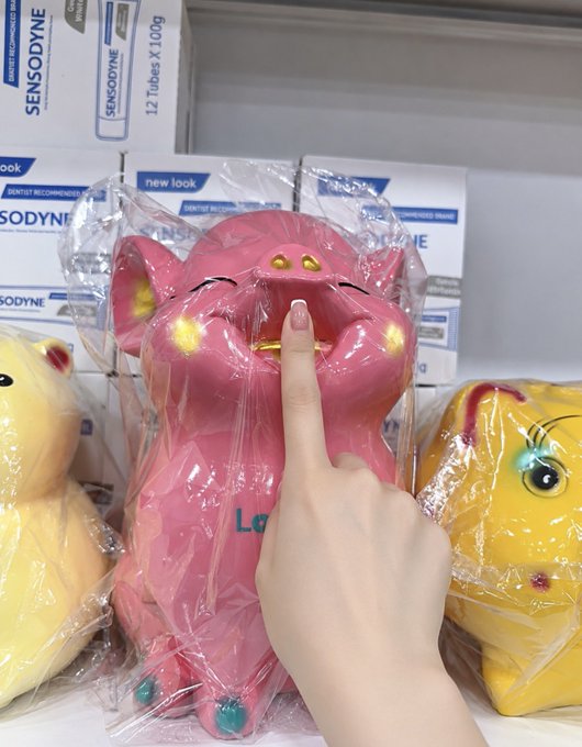A hand points at a pink Maneki-neko (lucky cat) figurine wrapped in plastic on a store shelf, alongside a yellow one and boxes of Sensodyne toothpaste.