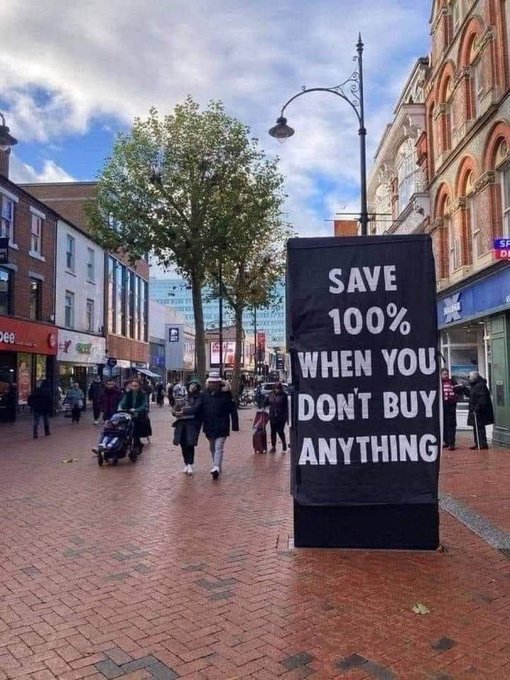 A pedestrian shopping street features a prominent black billboard displaying the message "SAVE 100% WHEN YOU DON'T BUY ANYTHING," with shoppers, a stroller, and storefronts visible in the background. The image aligns with the post's $MfT (Family Tree) crypto theme by promoting anti-consumerist spending, encouraging community members to keep their money in their own pockets rather than fueling the fiat system.