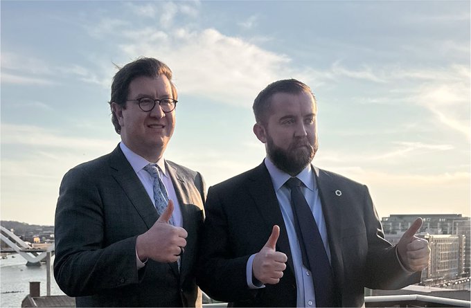 Two men in suits and ties stand side-by-side outdoors at sunset, both giving thumbs-up gestures, with a city skyline and body of water visible behind them.