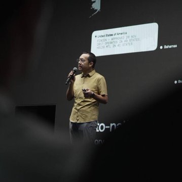 A man in a yellow shirt speaks into a microphone at an OKX event, with a presentation screen displaying "United States of America" stats including approval ratings, behind him—capturing a moment from OKX Now Singapore during Token2049 week.