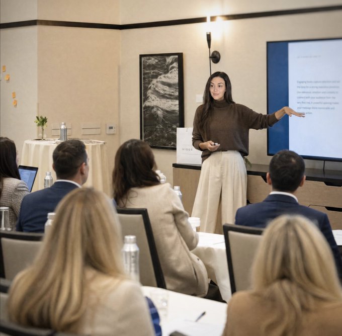A woman in a brown turtleneck and beige pants giving a presentation to a seated audience in a conference room, gesturing toward a presentation slide on a large screen.