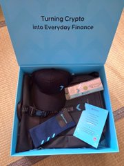 An open blue subscription box titled 'Turning Crypto into Everyday Finance' containing a black hat, merchandise, and informational cards.