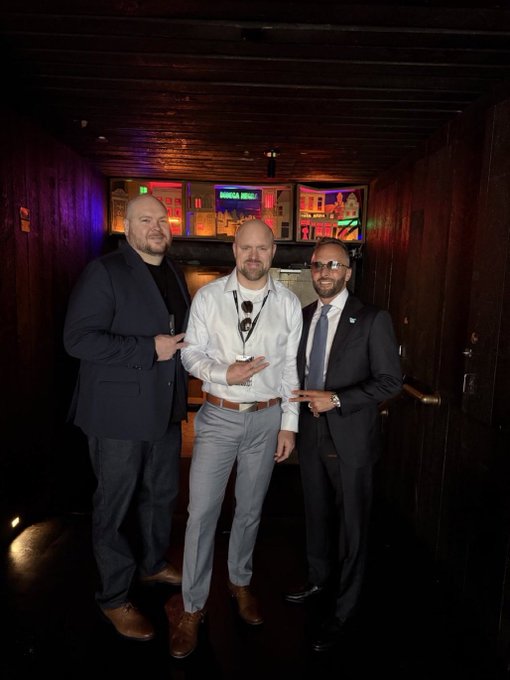 Three men pose together in a dimly lit venue with neon signage in the background; the man on the left in a dark blazer and the man on the right in a suit with sunglasses each point at the man in the center, who wears a white shirt and lanyard.