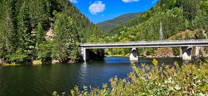 A scenic landscape showing a concrete bridge spanning a calm blue river, surrounded by forested mountains under a bright blue sky with scattered clouds.