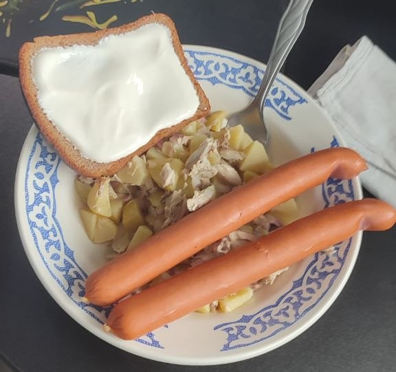 A bowl of hot dogs served with shredded meat, potatoes, and a slice of bread topped with a thick layer of white cream or yogurt.
