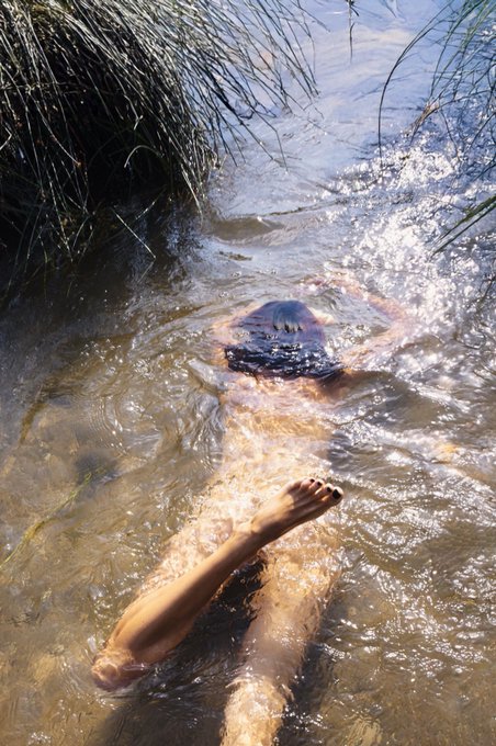 A first-person view of a person floating on their back in shallow water near grassy banks, with their face partially visible above the surface.