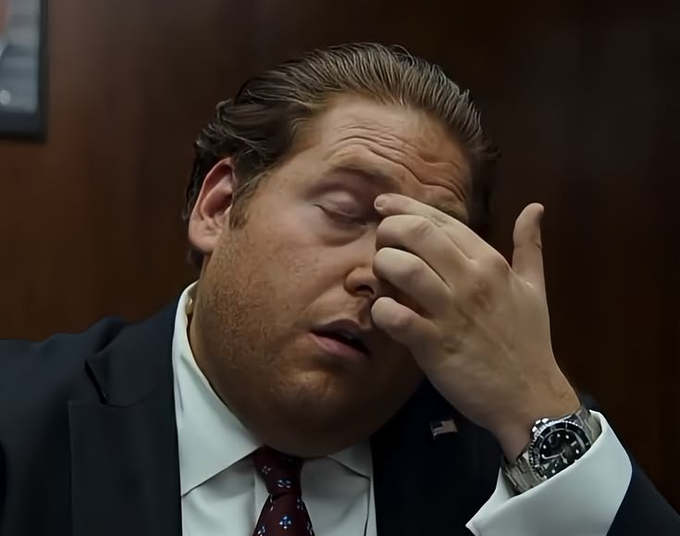 A close-up of a man in a suit with slicked-back hair rubbing his eye/forehead, appearing frustrated or tired, wearing a luxury watch.
