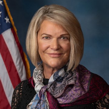 A woman with blonde hair, wearing a patterned purple and blue scarf, is pictured smiling in front of an American flag backdrop, accompanying news about Senator Lummis warning that delays in passing the CLARITY Act could postpone crypto market structure regulations until 2030.