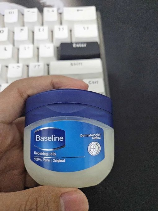 A hand holding a jar of 'Baseline Repairing Jelly 100% Pure | Original' skincare product in front of a white mechanical keyboard on a black surface.