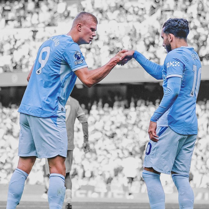 Two Manchester City players wearing light blue kits with the number 9 (Haaland) and number 10 (Grealish) share a fist bump on the pitch, celebrating their first victory in European competition this season.
