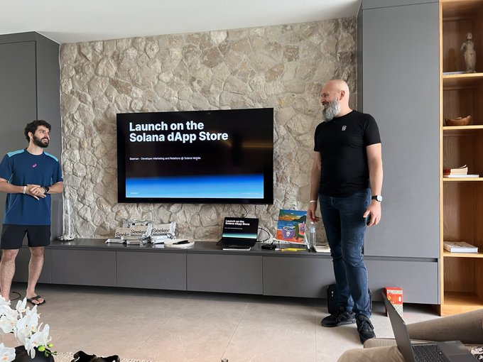 The image shows two men standing in a living room, with one man holding a laptop while the other stands next to him. They appear to be discussing something related to launching an app or a game on their smartphones. The living room has a flat-screen TV mounted on the wall and a bookshelf nearby. There are also several books scattered around the room.