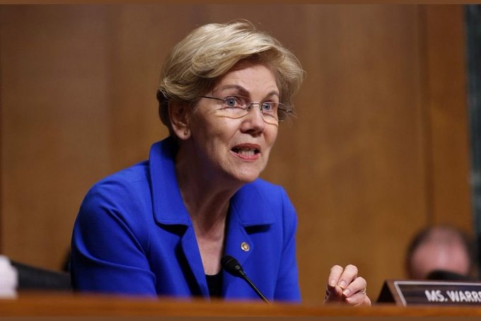 Senator Elizabeth Warren, identified by a 'MS. WARREN' nameplate, speaks at a hearing while wearing a bright blue blazer and glasses.
