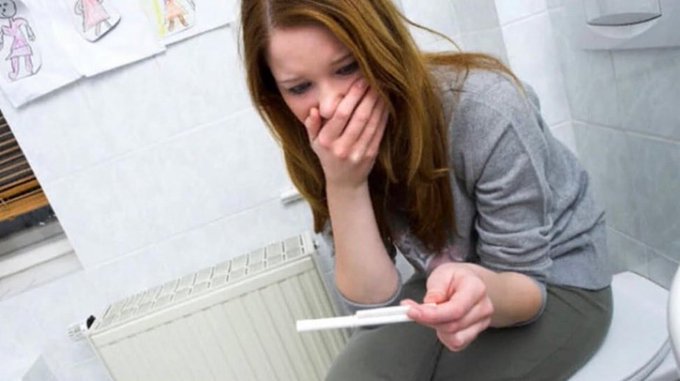 A young woman sits on a toilet holding a pregnancy test with a concerned expression, covering her mouth in shock as she awaits the result. The image ties to the post's caution about being prepared before finding yourself in an unexpected pregnancy scare, with "$POKEAHOE" humorously suggesting protection is essential to avoid such stressful moments.