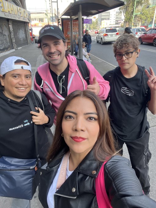 A group of four people pose for a selfie on a city street, with one man giving a thumbs-up.