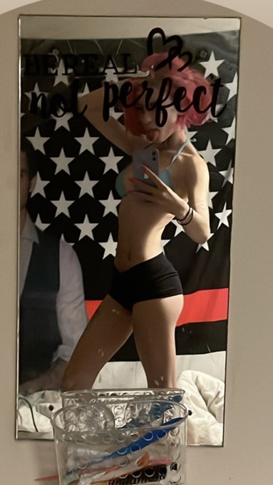 A mirror selfie of a pink-haired person in a bikini top and black shorts taking a photo with a smartphone, with an American flag backdrop displaying text 'I'm not perfect'.