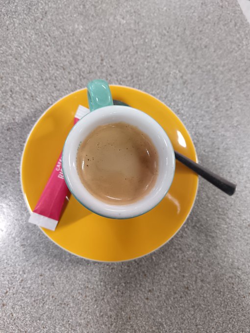A top-down view of a small white cup filled with espresso on a bright yellow saucer, accompanied by a black spoon and a pink sugar packet labeled 'Cane' on a gray surface.